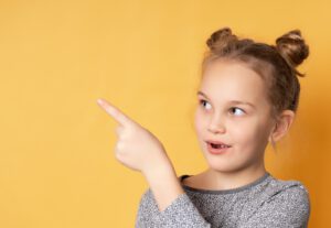 thoughtful little girl looking to the side and pointing finger to free space for text. Child poses against the background of a yellow wall in the studio. Banner. Advertising.